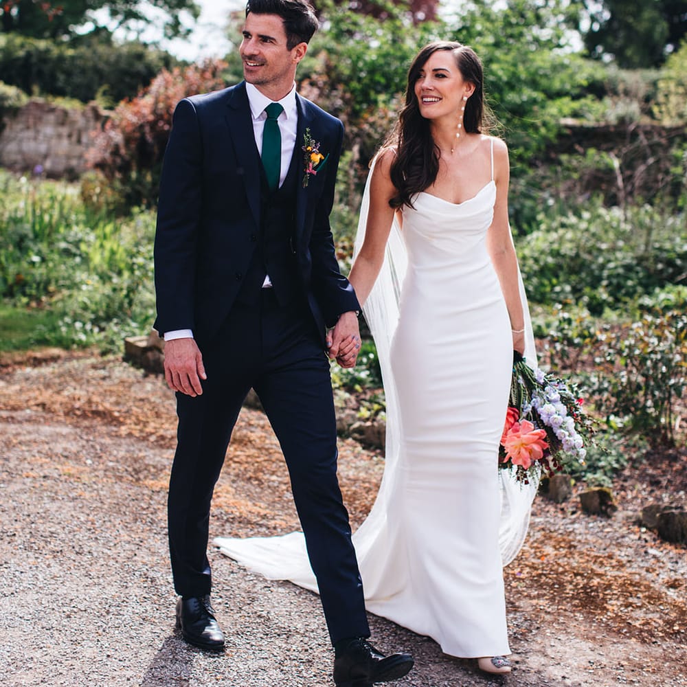 Groom in three piece suit with green tie and bright buttonhole walking with the bride in a fitted satin cowl neck Kyha wedding dress