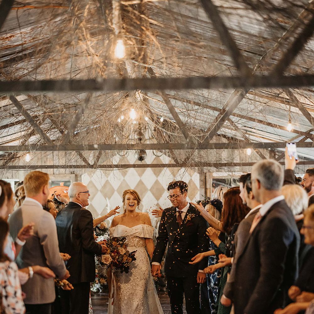 Bride in Love Spell wedding dress walks alongside her groom through colourful confetti exit 