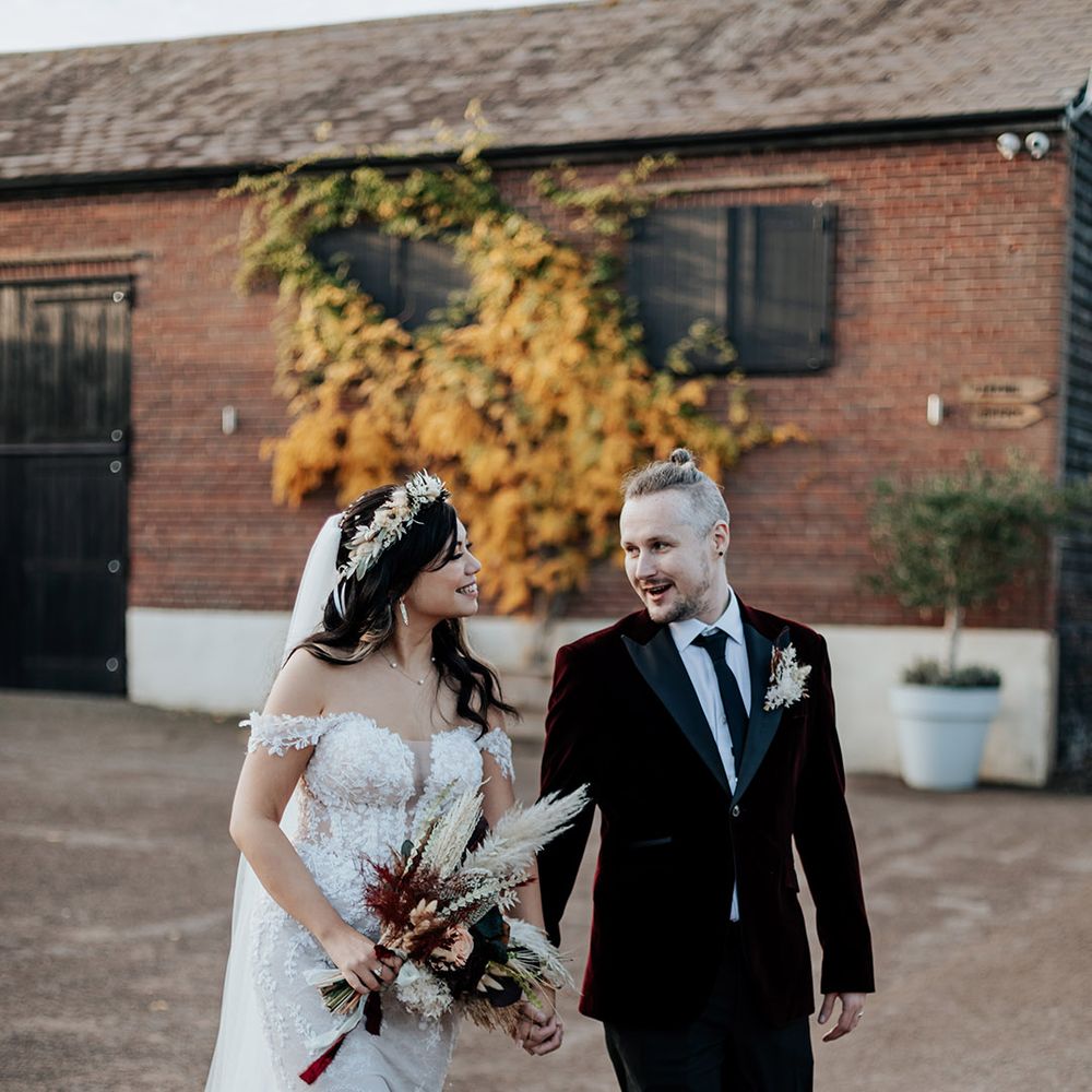 Milling Barn wedding with the bride in an off the shoulder lace wedding dress walking with the groom in a burgundy velvet wedding suit with dried flower bouquet, buttonhole and flower crown