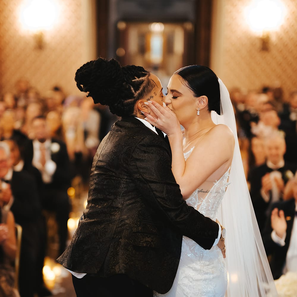 Bride in black snakeskin print tuxedo with black bowtie and white rose boutonniere kissing bride in sleeveless lace v neck Enzoani wedding dress with tulle finish and church length veil at black tie Grittleton House lesbian wedding 