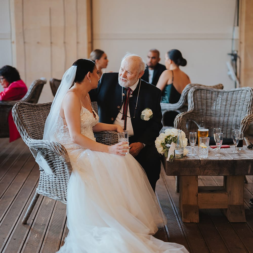 Bride sitting on a wicker chair at Grittleton House wedding venue in lace sleeveless v neck Enzoani wedding dress with tulle finish and church length veil with 3d appliqué embroidered flowers chatting with wedding guest 