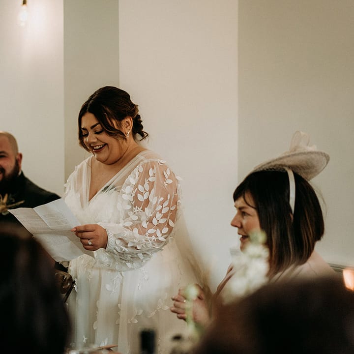 Bride in long sleeve lace wedding dress by All Who Wonder reading out her bridal speech 