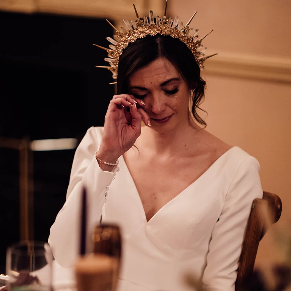Bride in long sleeve wedding dress wipes tears during the wedding ceremony as she feels emotional wearing a gold bride crown 