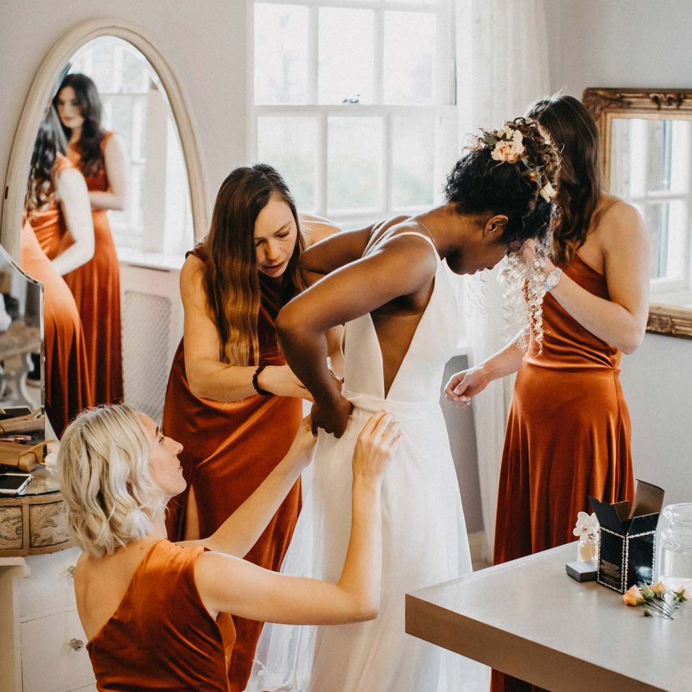Bridesmaids in one shoulder burnt orange bridesmaid dresses helping bride into her v neck sleeveless wedding dress with 3D floral applique train at Brinsop Court wedding