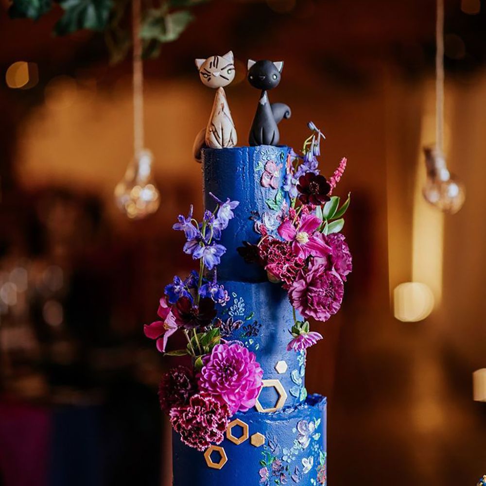 Extravagant dark blue and purple wedding cake with flowers, cat cake toppers and gold hexagonal detailing 