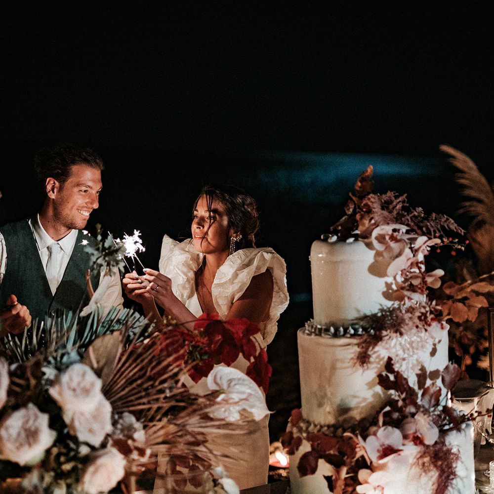 Outdoor wedding reception on the beach in Italy with giant wedding cake with dried flower decor and bride and groom holding sparkles 