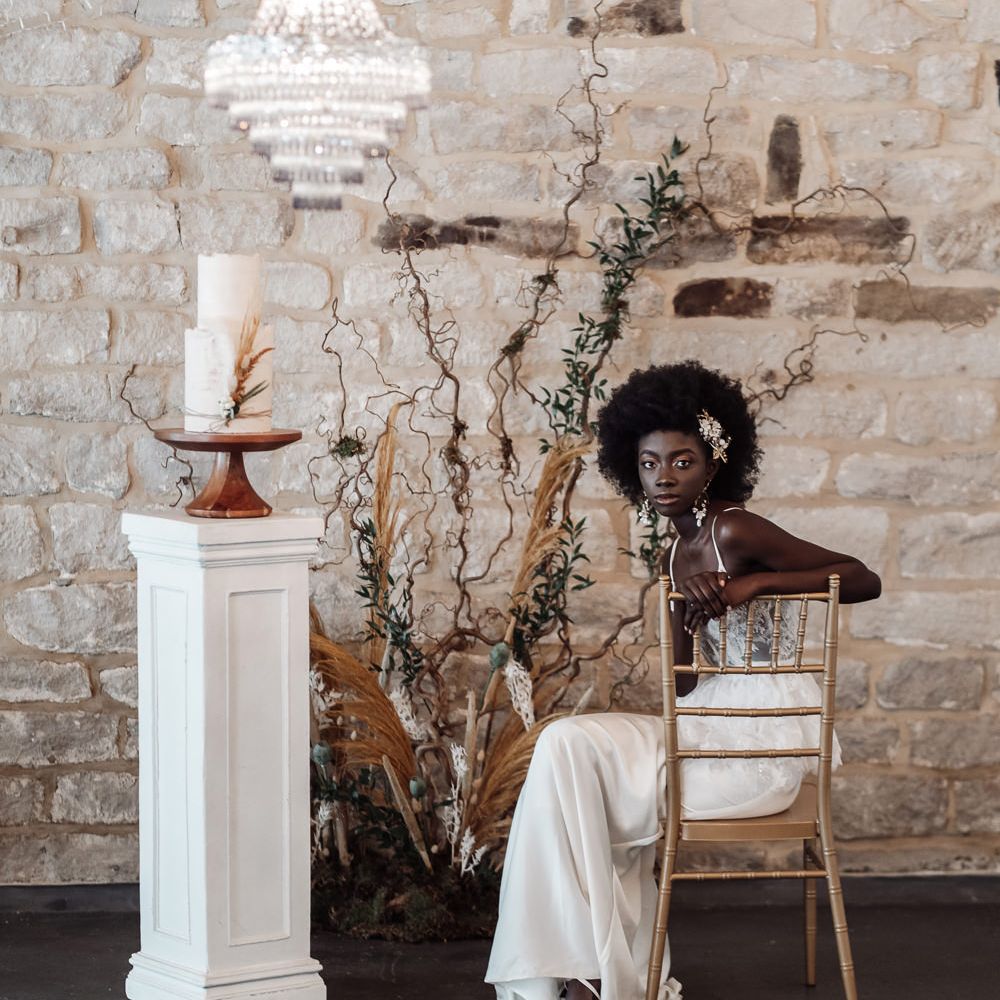 Black bride in a fitted wedding dress sitting on a chair at Ponden Mill with chandelier light installation, two tier wedding cake on a plinth and dried flower and foliage floral arrangement 