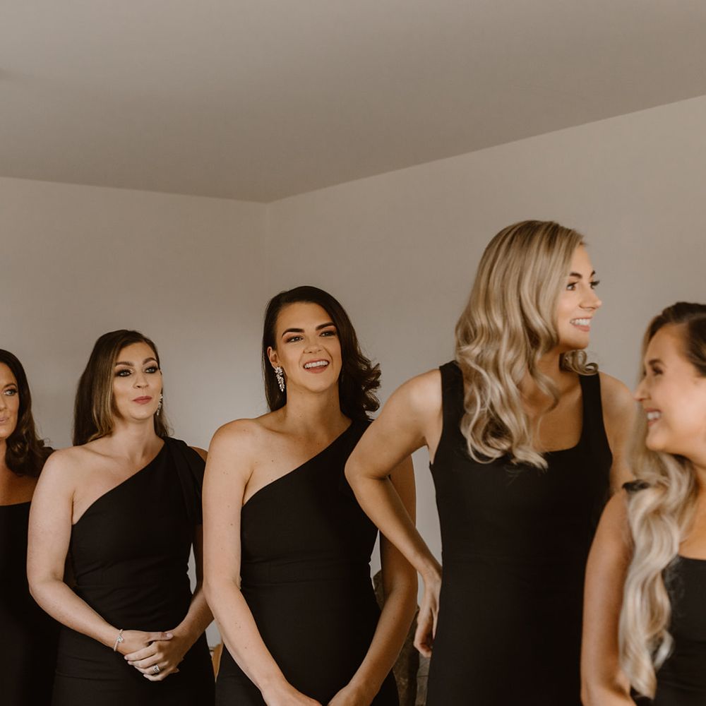Bridesmaids with curled hair and black bridesmaids dresses stand with father of the bride in black tuxedo before the wedding