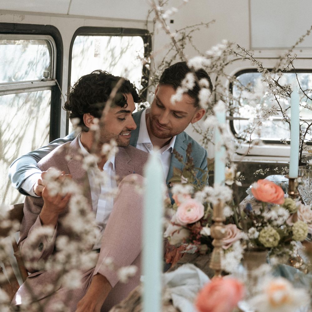 Grooms sit together surrounded by rustic floral decor and candlesticks in brass holders 