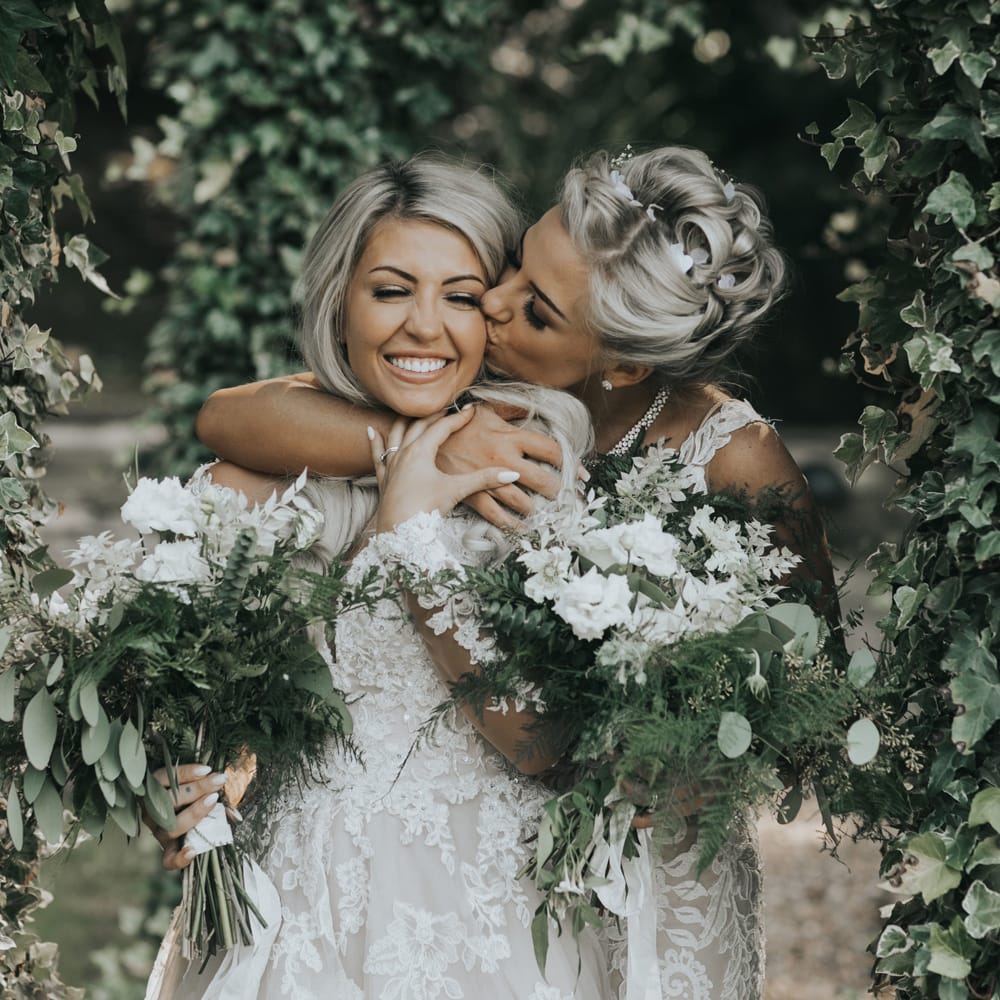 Two brides in white dresses holding white and green bouquets embrace under trees at crab & lobster fairytale wedding. 
