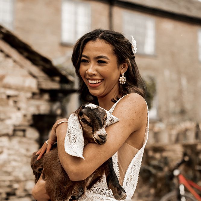 Black bride in a fitted wedding dress with straps holding a goat at their farm wedding venue