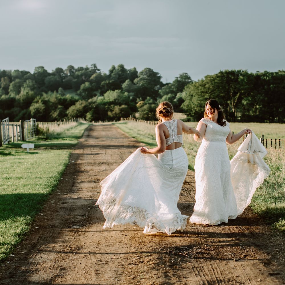 Brides walk through countryside on their wedding day as they lift their skirts and dance with one another
