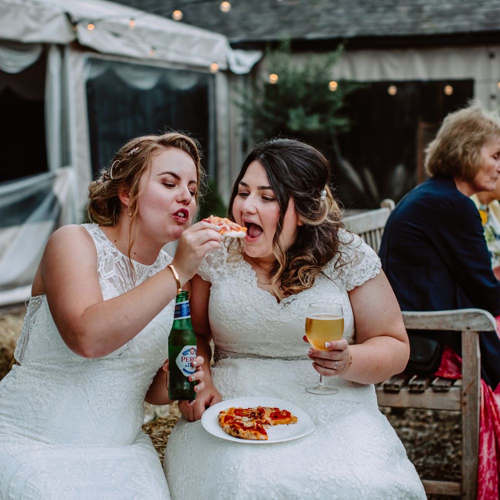 Bride feeds her bride pizza on the day of their wedding as they sit outside at Park Farm