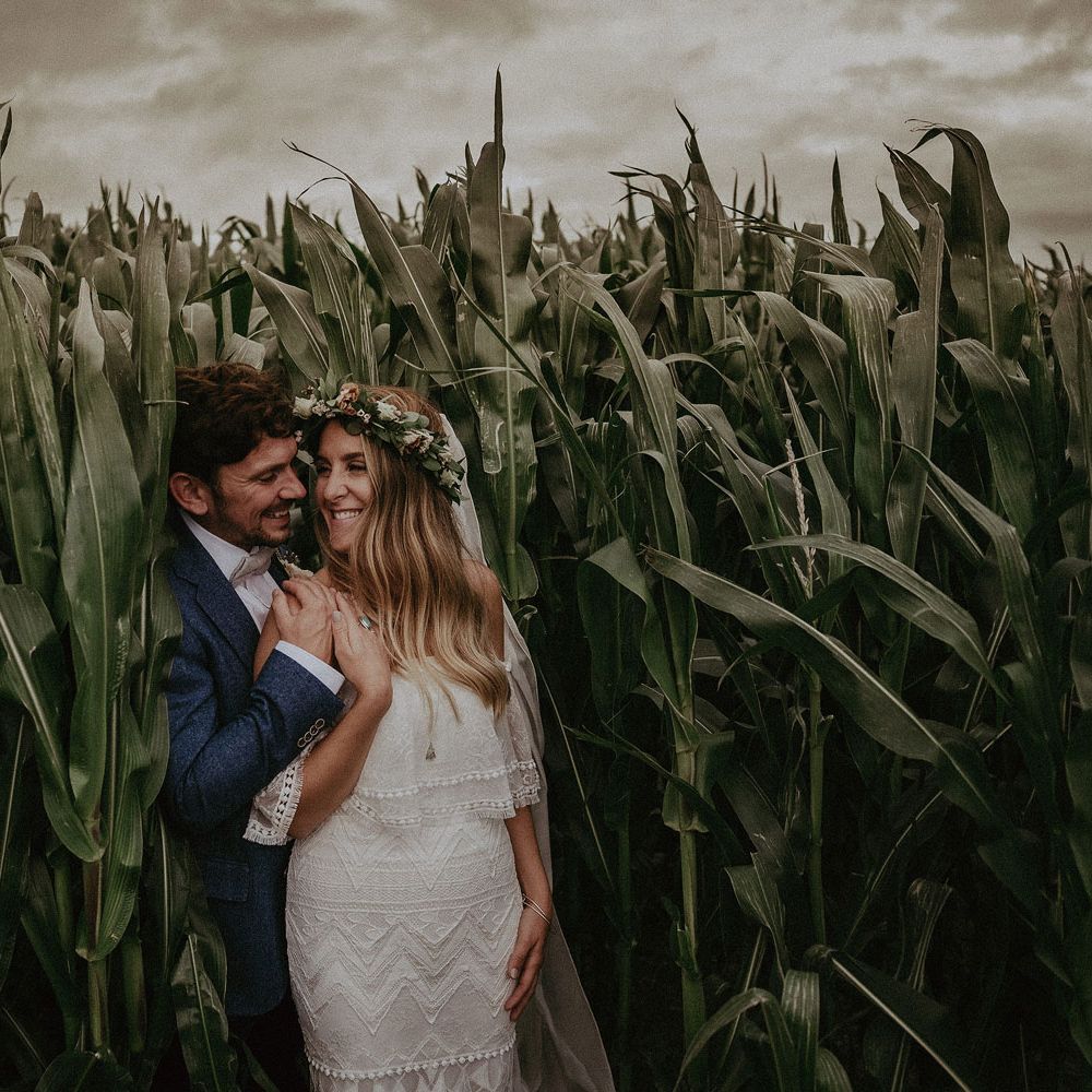 Bride in Grace Loves Lace bardot wedding dress, flower crown and veil stands with groom in blue suit jacket in corn field after Isle of Wight wedding with macrame wedding decor
