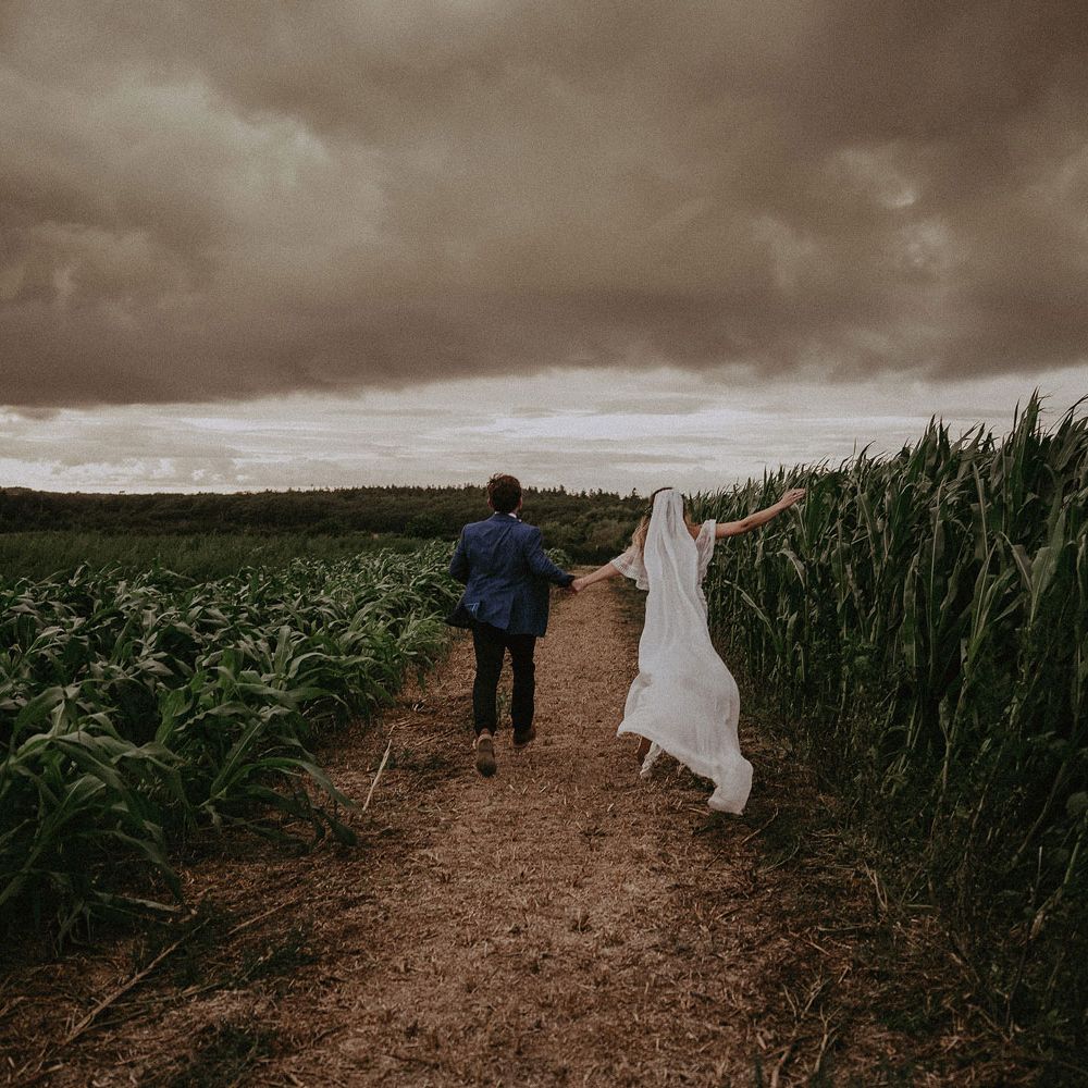 Bride in Grace Loves Lace wedding dress and veil runs through corn field holding hands with groom in blue suit jacket at Isle of Wight wedding with macrame wedding decor