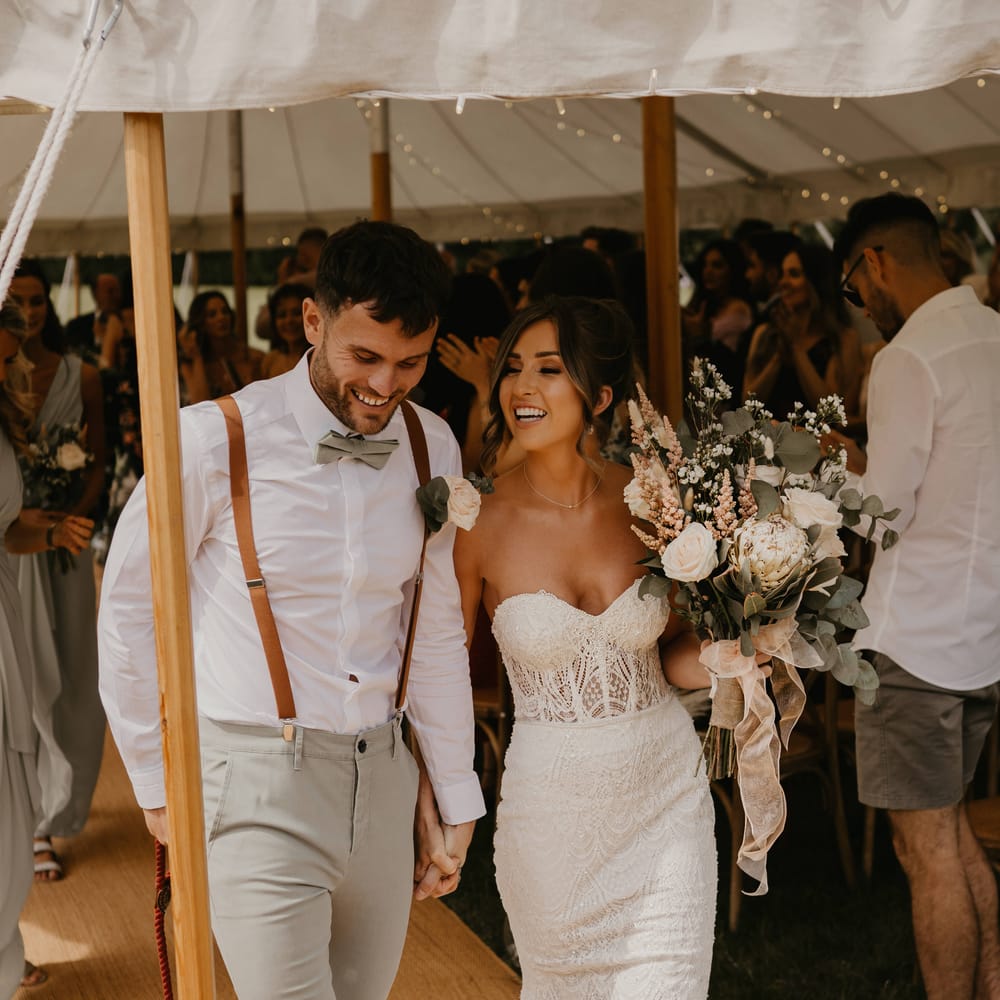 Bride & groom hold hands as they walk through marquee and bride holds her floral bouquet | Mark Bamforth Photography