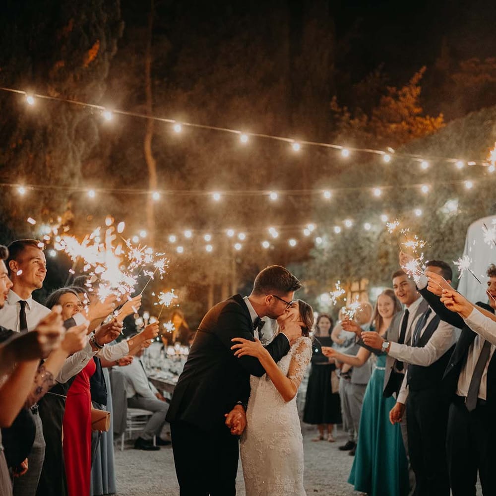 Bride & groom kiss as wedding guests hold sparklers and fairy lit canopy hangs above them | Royal Studio