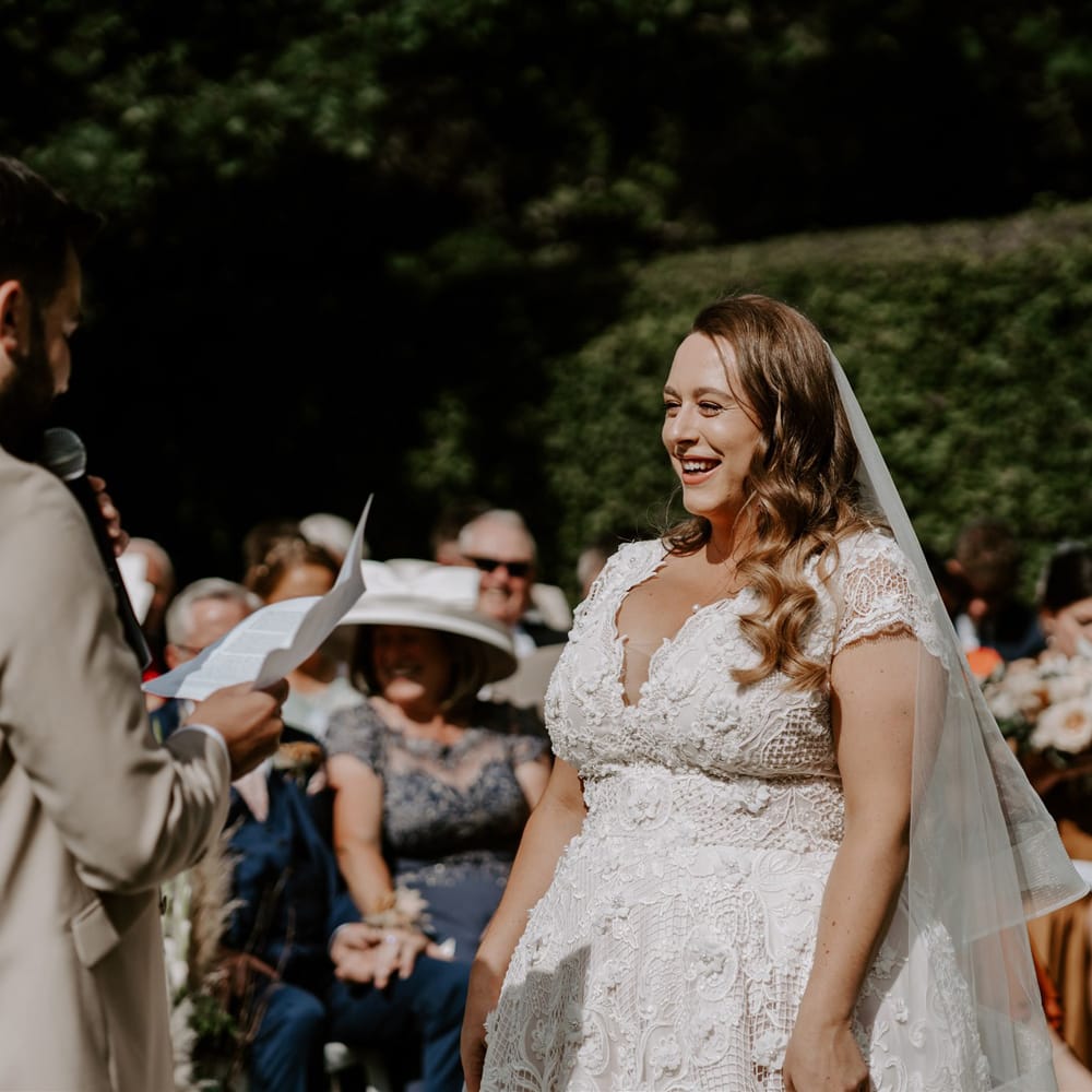 Bride in homemade lace wedding dress and veil laughs as groom in beige suit reads out vows during DIY garden wedding ceremony in Bedfordshire