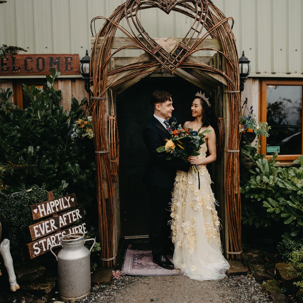 Groom in dark suit stands in doorway with bride in strapless feathered wedding dress and headpiece for Wellbeing Farm wedding in Lancashire 