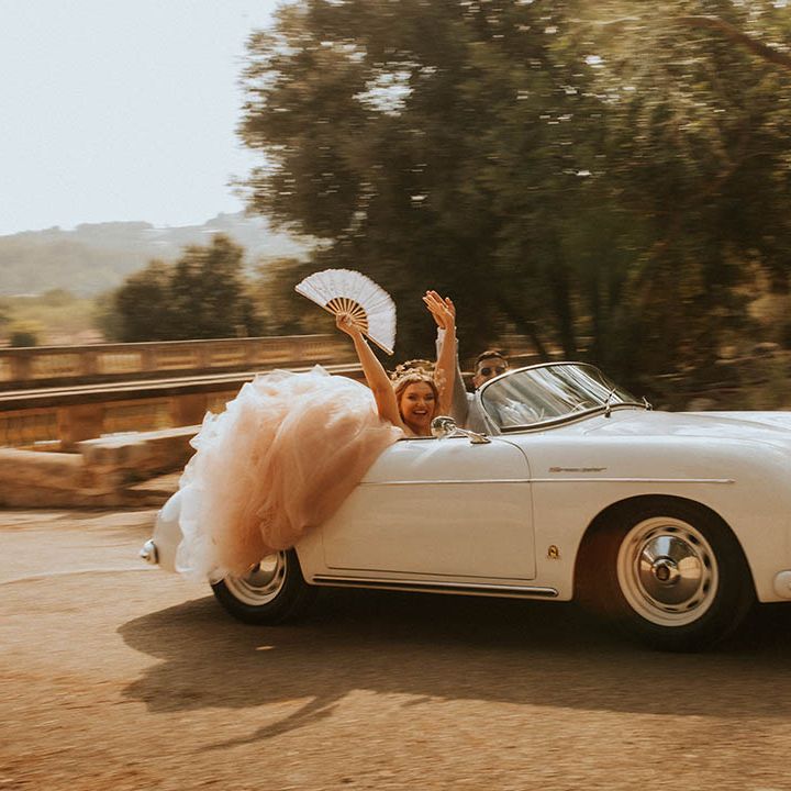 Bride & groom smile and wave as they drive in vintage car