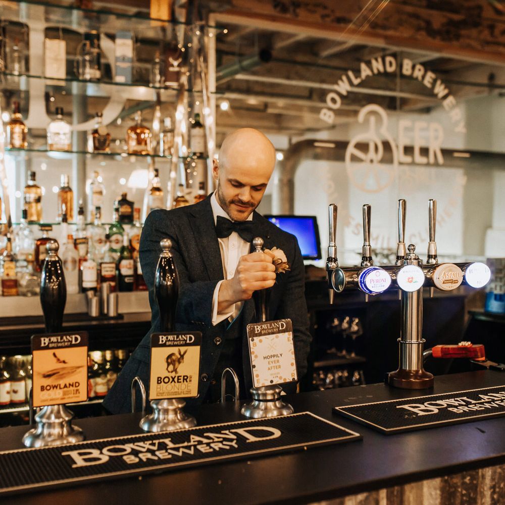 Groom in navy tweed suit and bow tie pours himself an ale at wedding reception