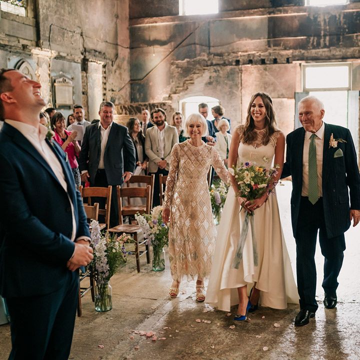 Bride is walked up the aisle in lacy two piece wedding top and high low skirt with blue wedding shoes and wildflower bouquet