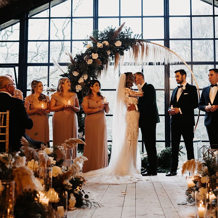 Bride and groom share a kiss after humanist wedding ceremony with bridal party and groomsman standing to the side holding candles 