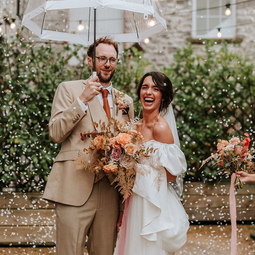 Bride in white puff sleeve wedding dress and Loeffler Randall shoes with groom in beige three piece suit holding clear umbrella as confetti is thrown 