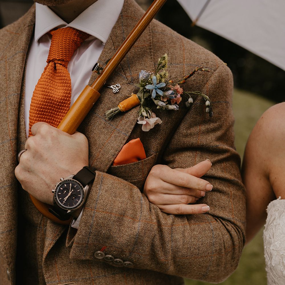 Bride links arms with the groom in brown suit with orange tie and colourful buttonhole with black watch