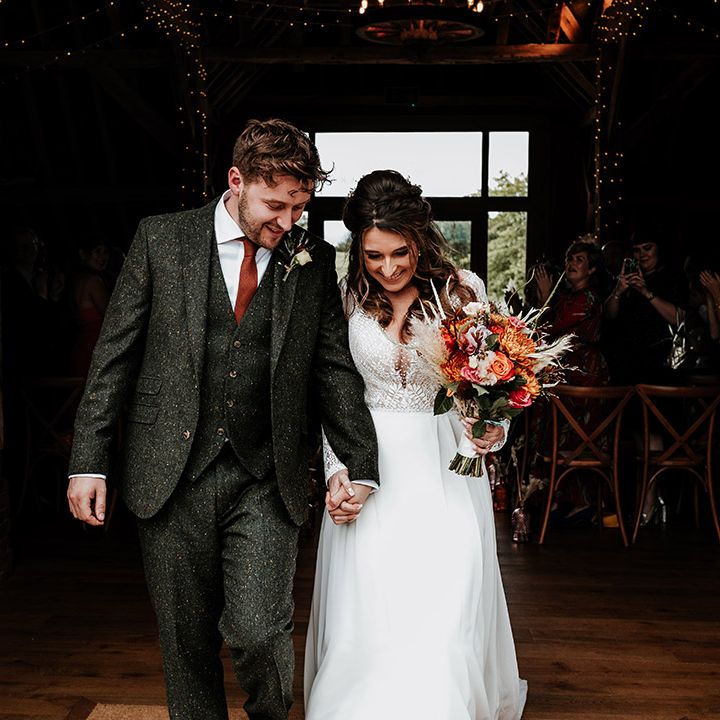 Groom in green wool three piece suit with brown tie exits ceremony holding hands with bride in lace top wedding dress