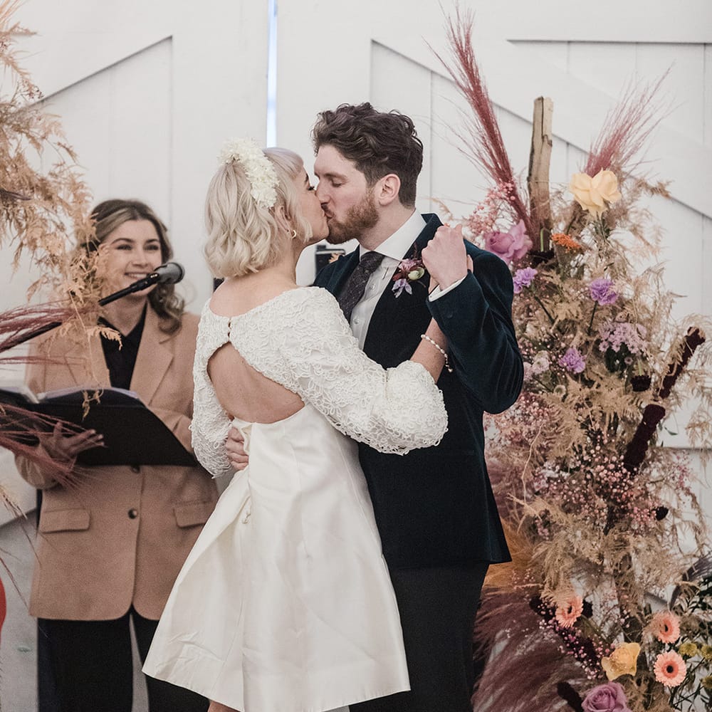 Bride in short wedding dress with platform black block heels kissing the groom in velvet suit next to dried wedding flowers