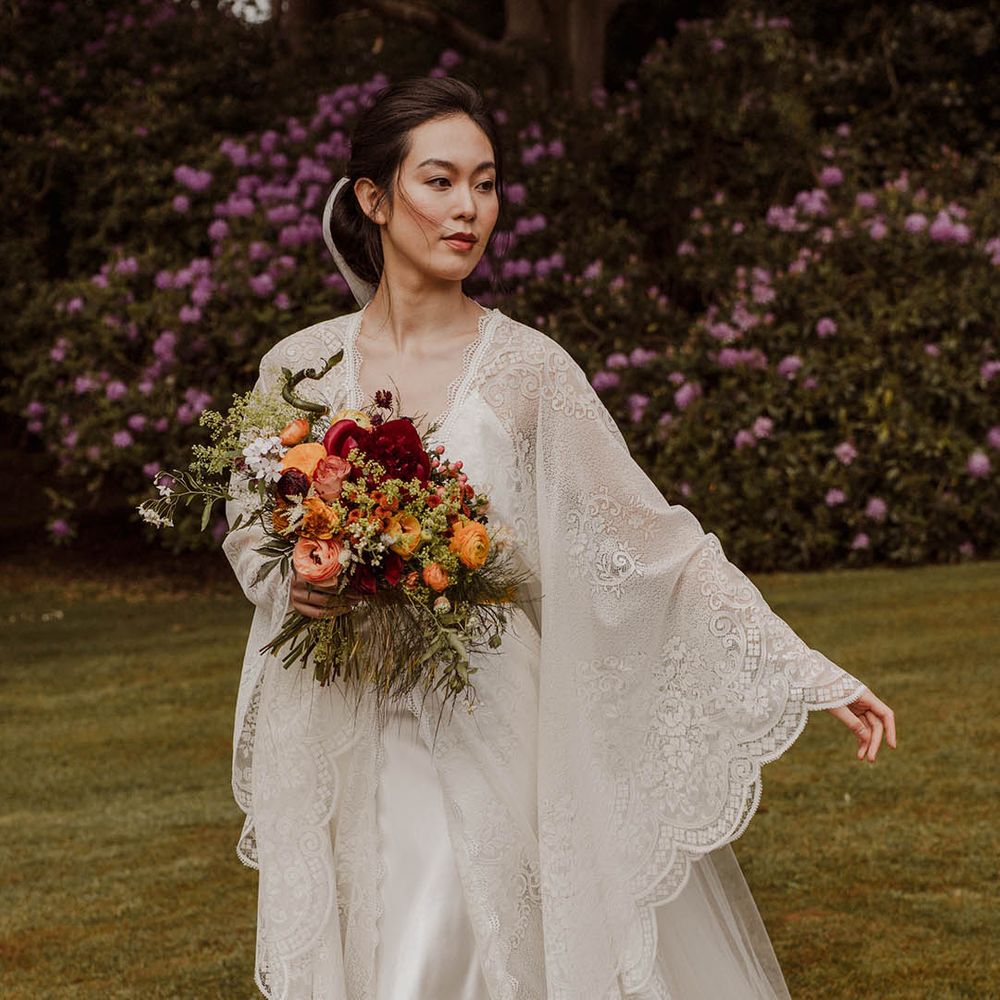 East Asian bride in a lace Kimono style wedding dress with long sleeves holding a red, yellow and orange wedding bouquet 
