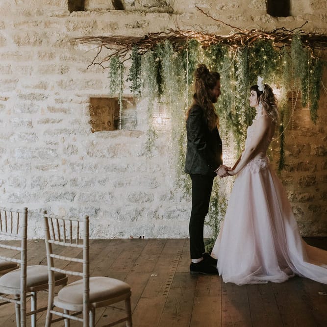 Bride in a pink wedding dress and groom in a black suit exchanging vows inside Hooton Pagnell's wedding ceremony room with a fairy light, foliage and twig backdrop
