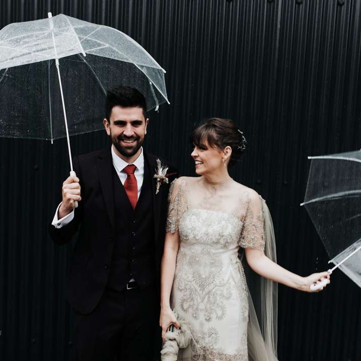 Bride in homemade sage tinted quarter length sleeve wedding dress with homemade bouquet and peacock feathers and groom in dark suit with red tie holding clear umbrellas