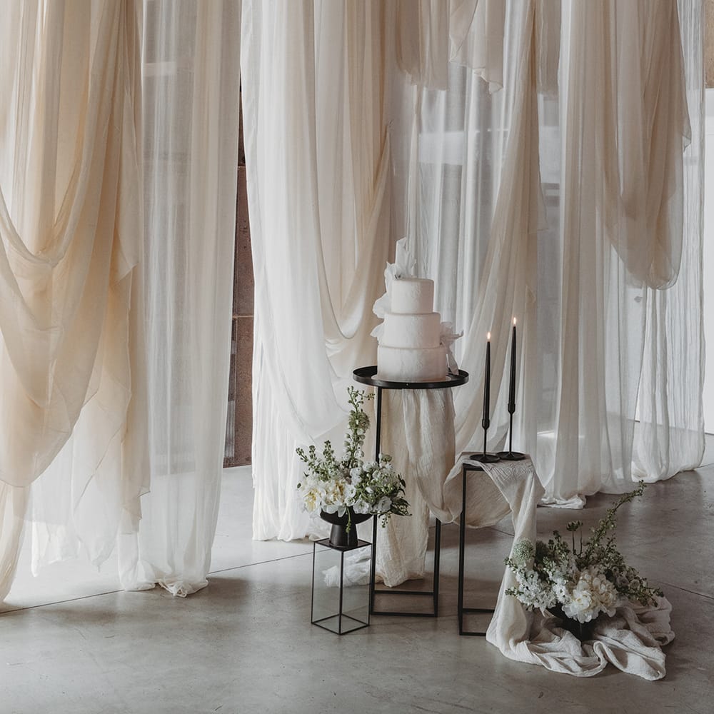 White wedding drape backdrop with a white iced wedding cake with black taper candles and dark metal decorated with white flowers 