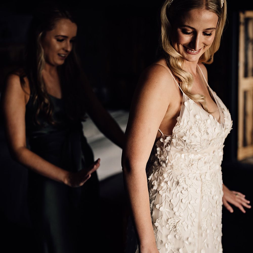 Bridesmaid in green satin dress helps the bride into her Alexandra Grecco Lana wedding dress with a pretty pearl headband 