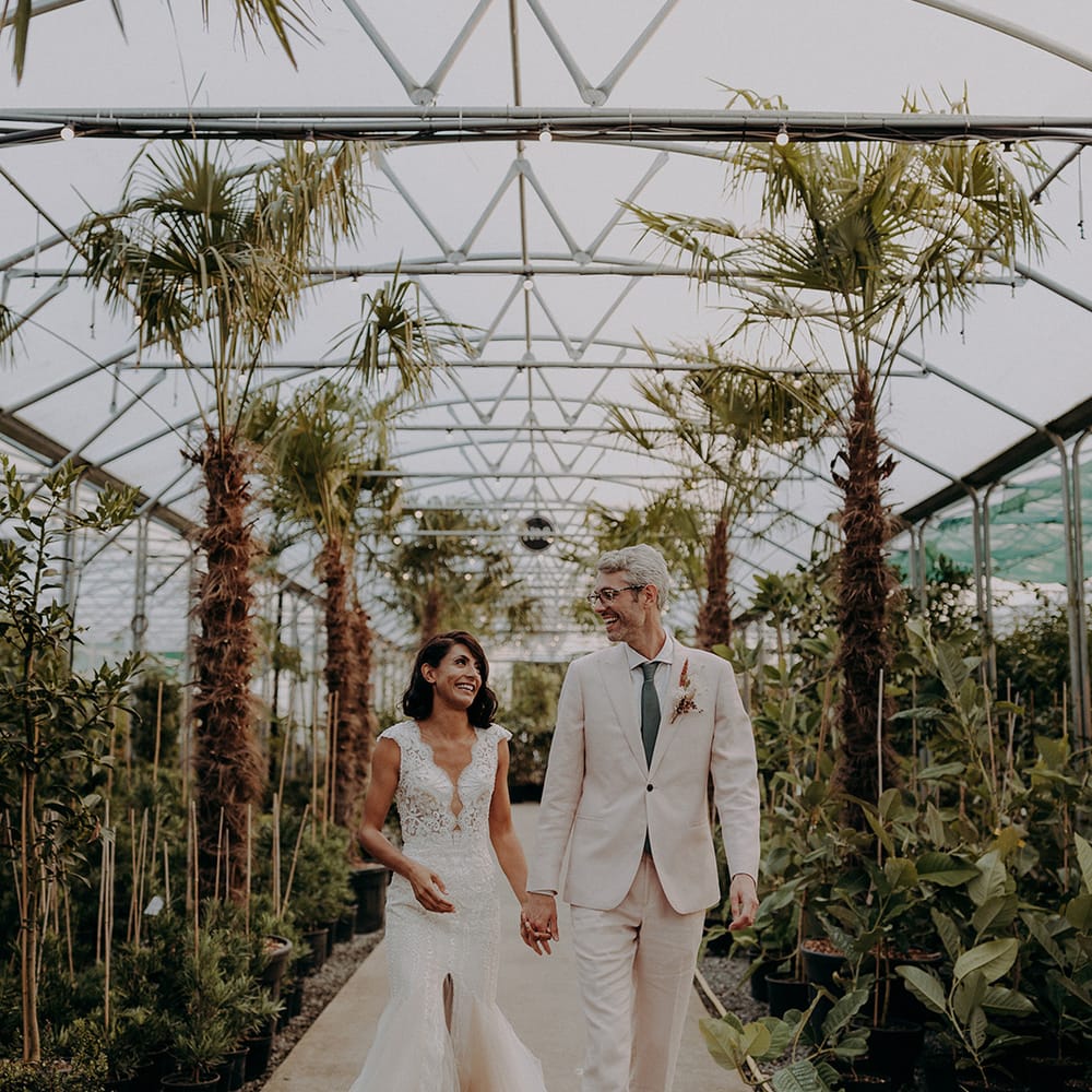 Bride in lace and tulle dress and groom in light pink suit hold hands whilst walking through plants and trees