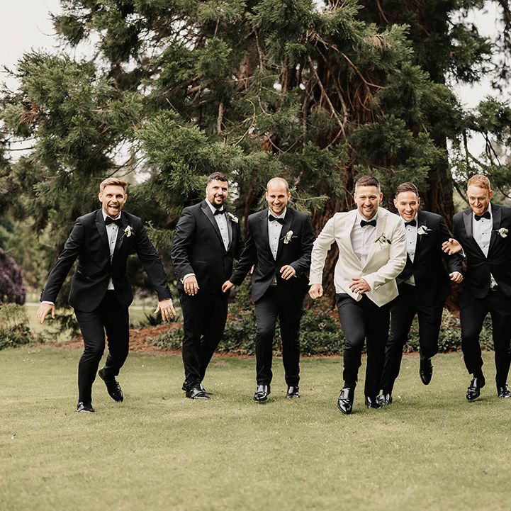Groom in a white suit jacket and black trousers running with the groomsmen in black tie 