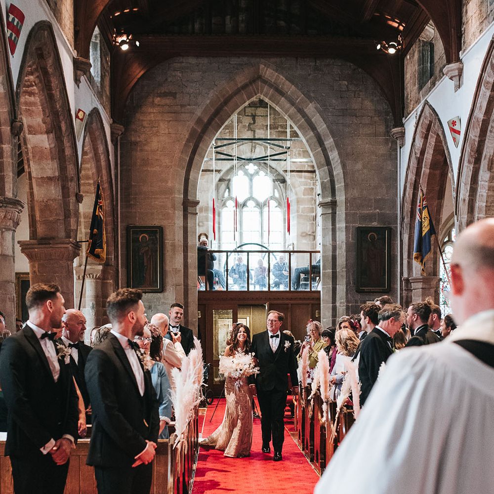 Father of the bride in a black tuxedo walks the bride down the aisle on a red carpet to the groom waiting at the end also in black tie 