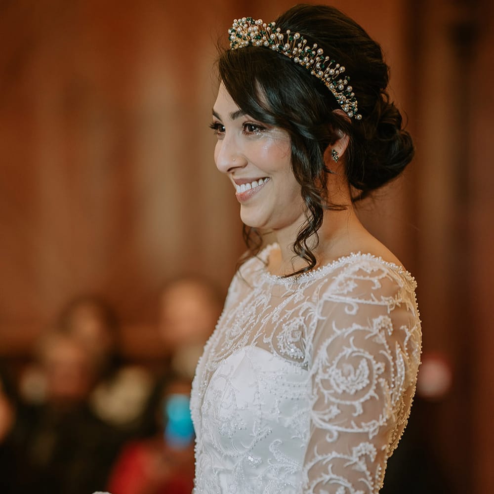 Bride in princess wedding dress with long lace sleeves and gold, pearl and emerald bridal crown standing at the alter