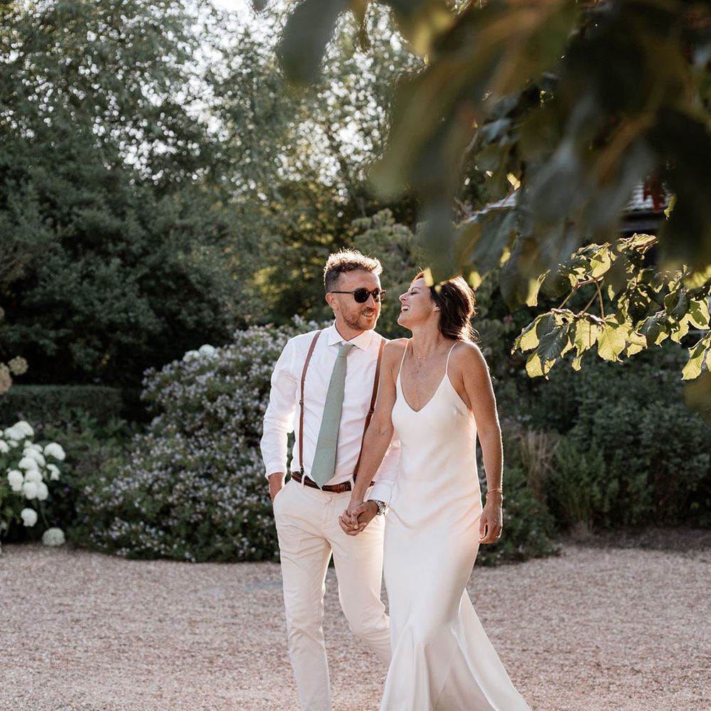 Bride in fitted silk and simple Grace Loves Lace wedding dress walks alongside her groom in sage green tie and braces