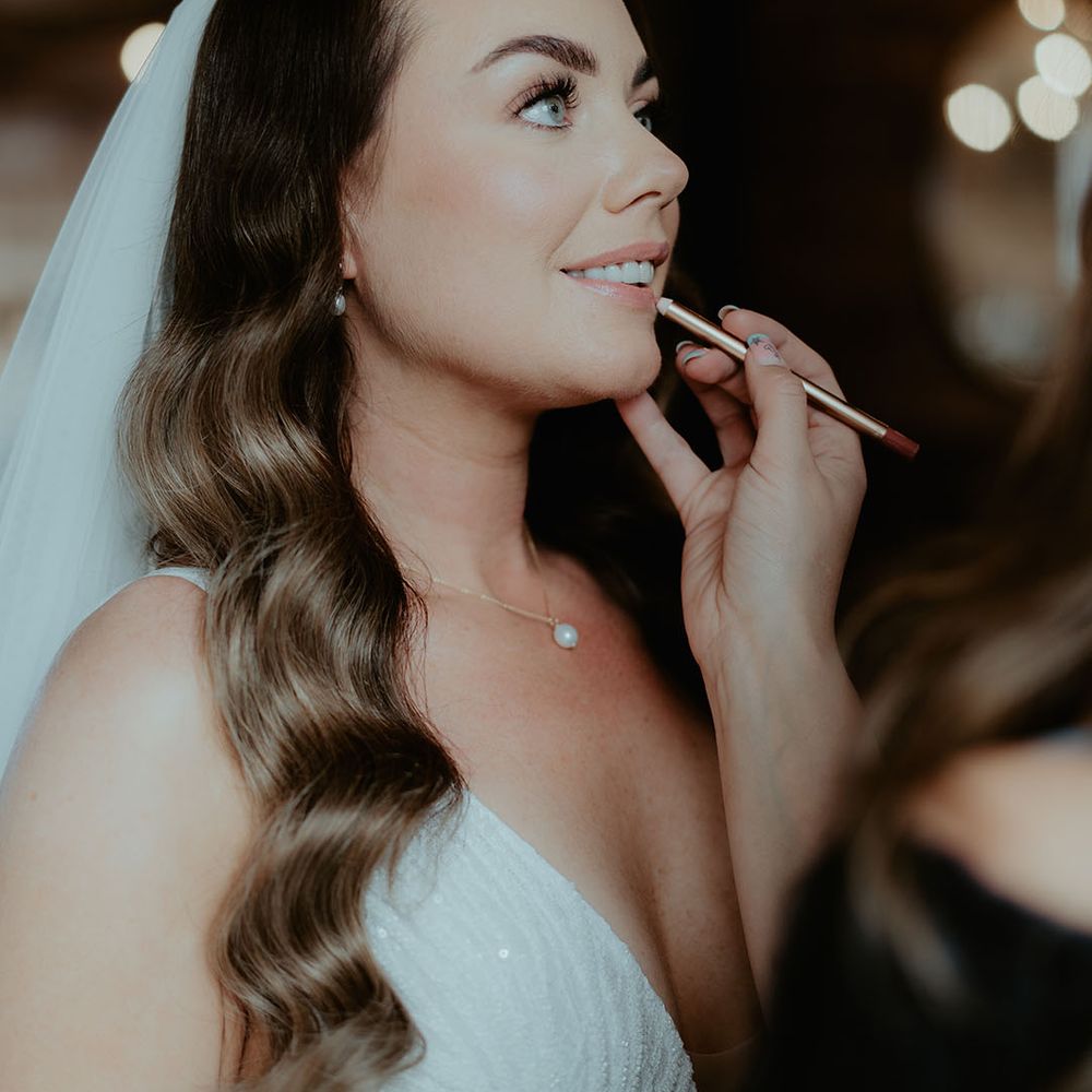 Bride with her brown hair in soft Hollywood waves stands in her embellished wedding dress and veils stand getting her makeup done with Charlotte Tilbury products 