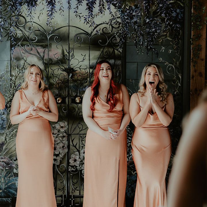 The bridesmaids in matching satin cowl neck peachy orange bridesmaid dresses smile as they get their first look at the bride 