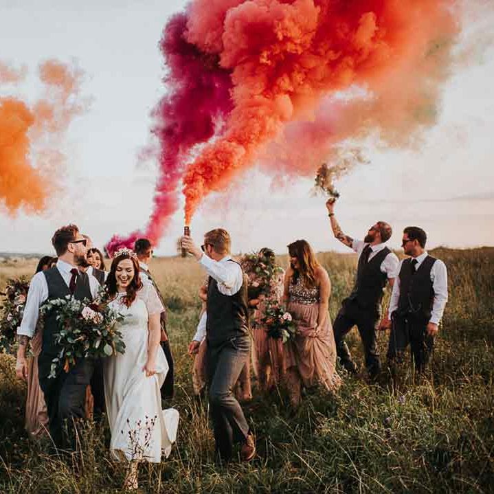 Bride in sheer lace sleeve wedding dress and groom in grey waistcoat and sunglasses walking with wedding party doing colourful orange and pink smoke bomb wedding photos 