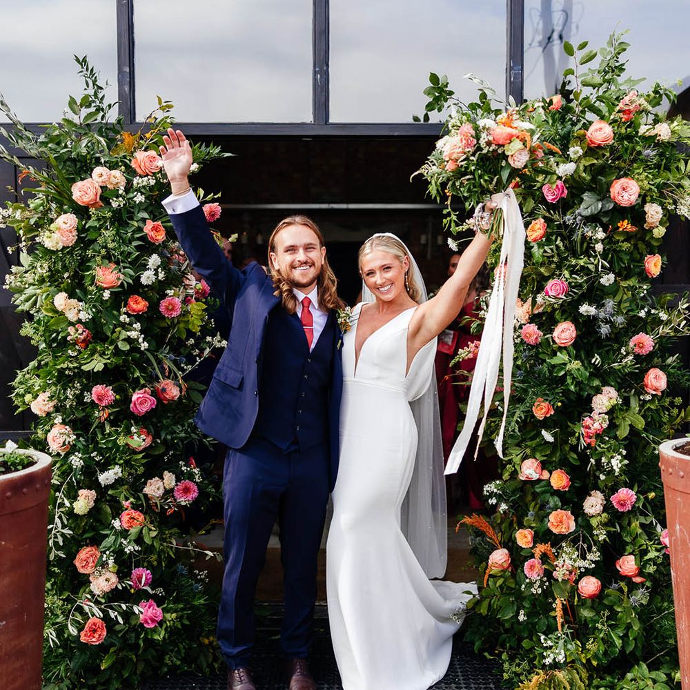 Bride in a fitted wedding dress with pearl earrings with the groom in blue three piece suit celebrating with the bride next to the flower column arrangements