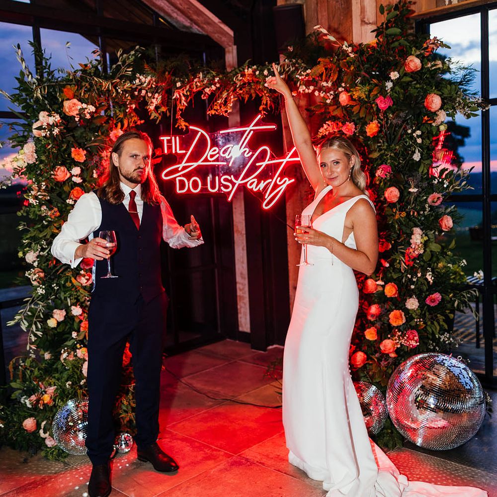 The bride and groom pose next to their red neon wedding sign reading 'til death do us party' with flower column decor 