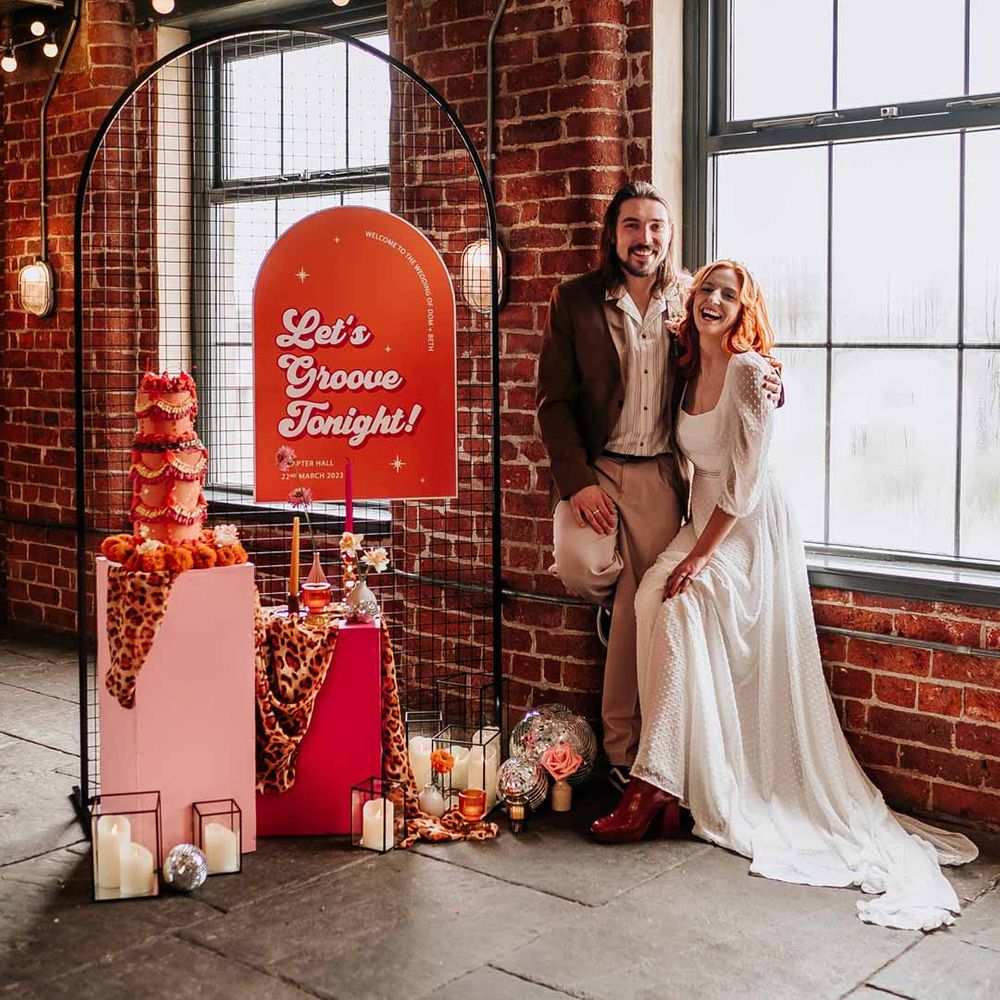 Bride in three quarter length puff sheer sleeve wedding dress with delicate polka dot overlay and square neck posing with groom in brown blazer, white shirt, grey suit trousers and vans shoes next to retro wedding decor