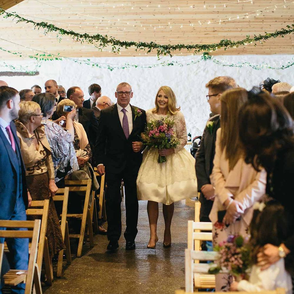 Bride in short long sleeve wedding dress walking down the aisle at Dalduff Luxury Barn Weddings with father of the bride 