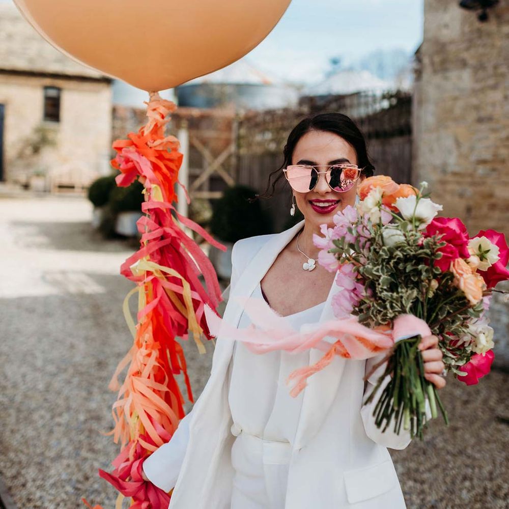 Bride in white bridal suit with rose gold wedding sunglasses holding large mixed seasonal flower bridal bouquet and large balloon with wedding streamers attached