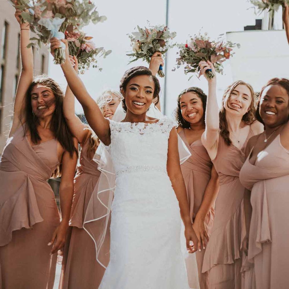 Bride wearing short sleeve boat neck lace wedding dress standing with bridesmaids in neutral ruffle bridesmaid dresses 